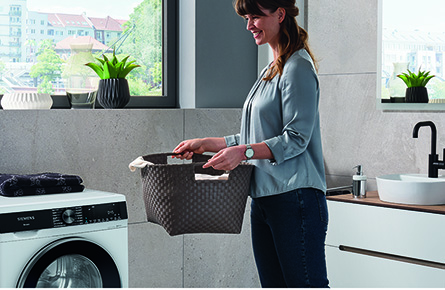 Frau hält einen Wäschekorb und steht vor einer Waschmaschine